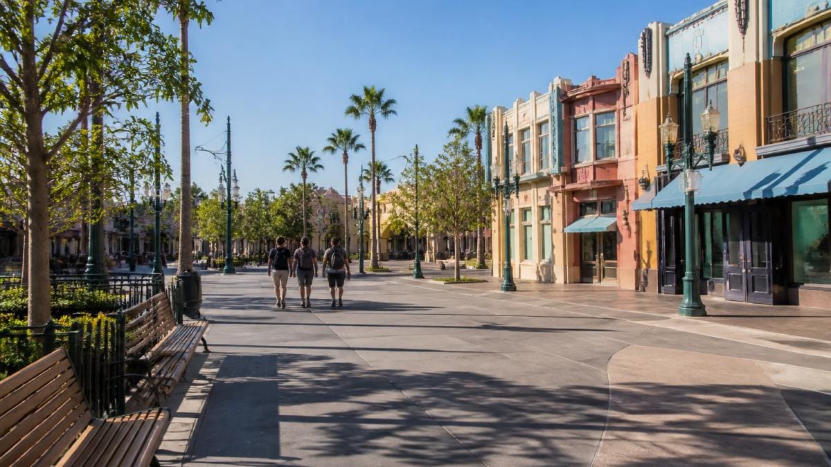 A wide shot of a mostly empty theme park walkway in the morning light, with a few visitors strolling past colorful storefronts and no visible ride queues
